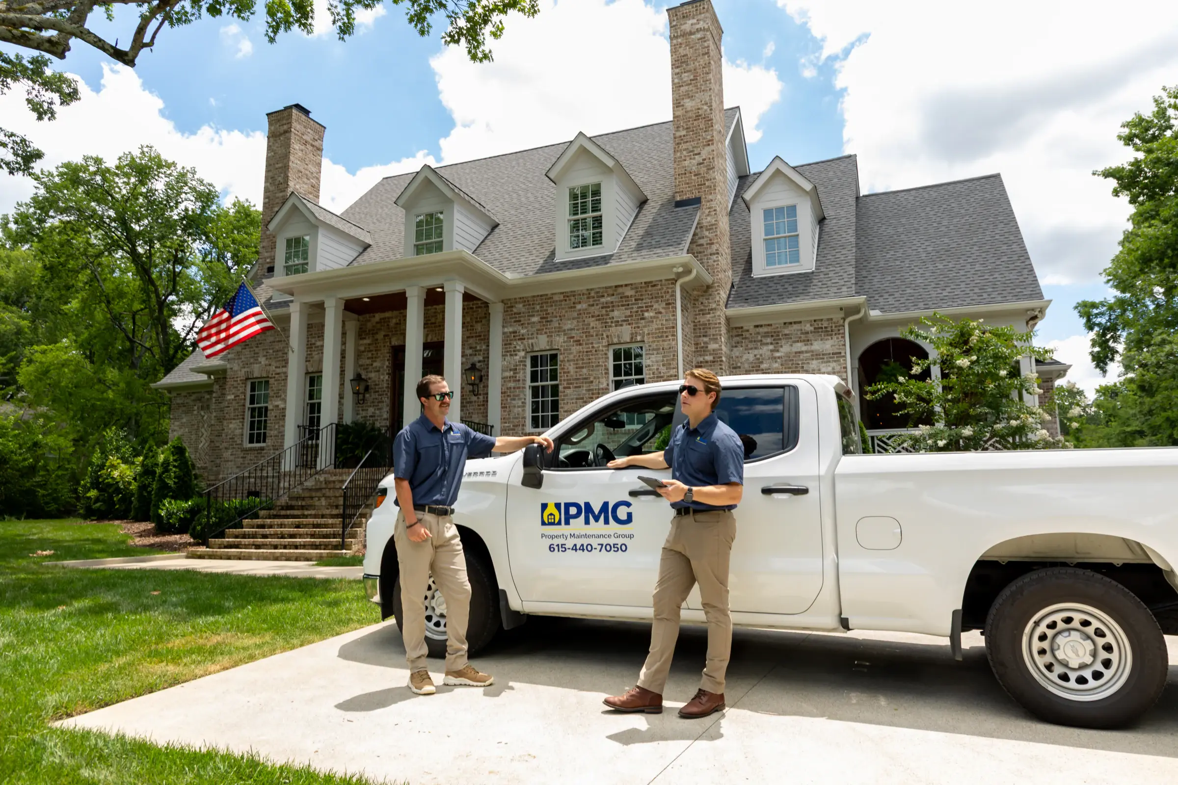 Two PMG technicians at the company truck in the driveway of a brick home